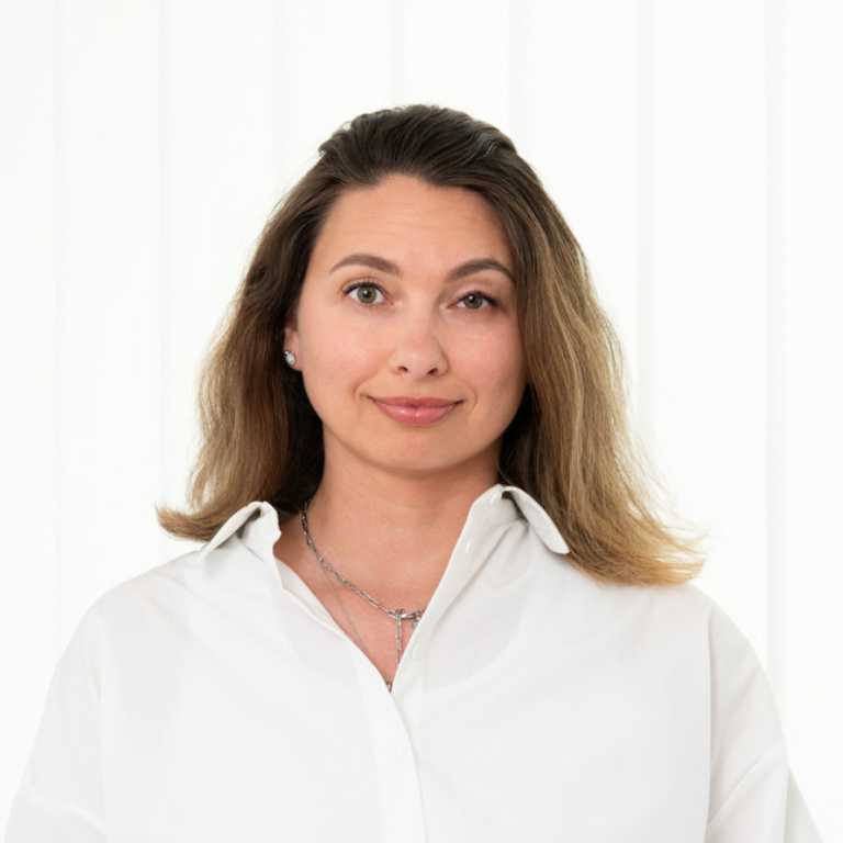 Frau mit schulterlangen, lockigen Haaren und einem freundlichen Lächeln, trägt ein weißes Hemd und steht vor einer hellen, strukturierten Wand.