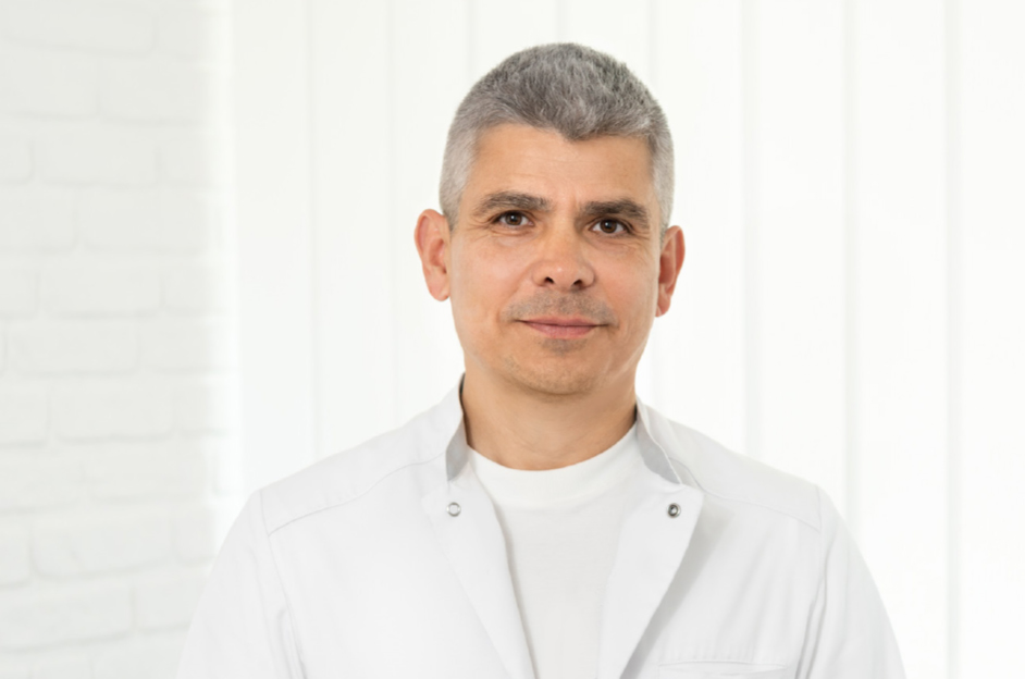 Mann mittleren Alters in medizinischer Kleidung, lächelnd vor hellem, neutralem Hintergrund, ideal für medizinische oder gesundheitliche Themen.