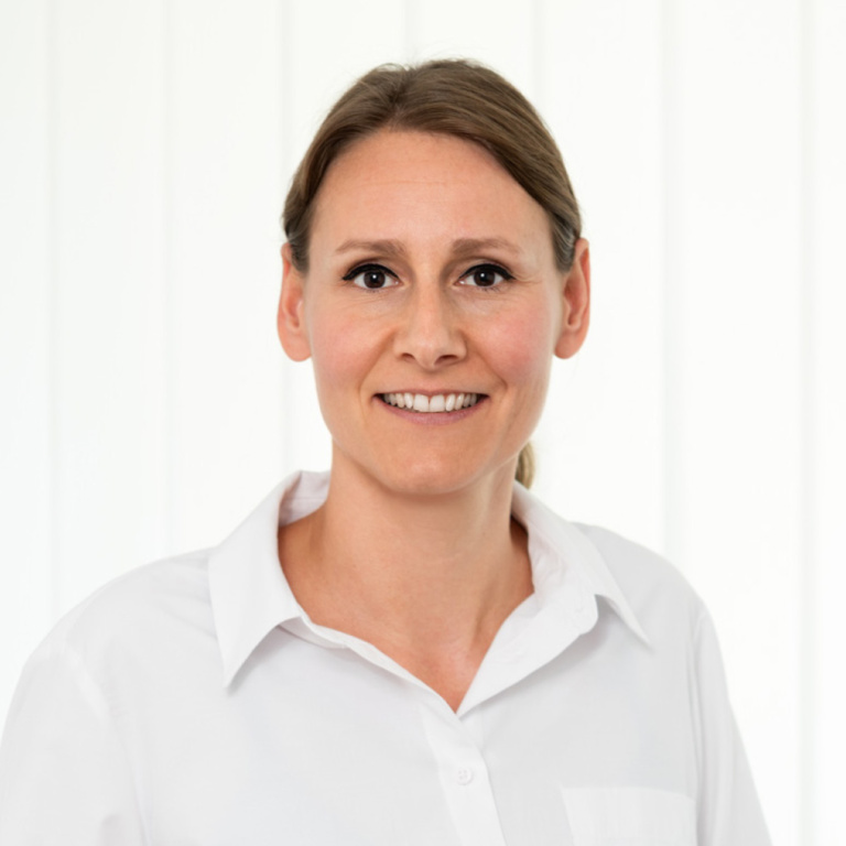 Portrait einer Frau mit freundlichem Lächeln, trägt ein weißes Hemd, vor einem hellen, neutralen Hintergrund.