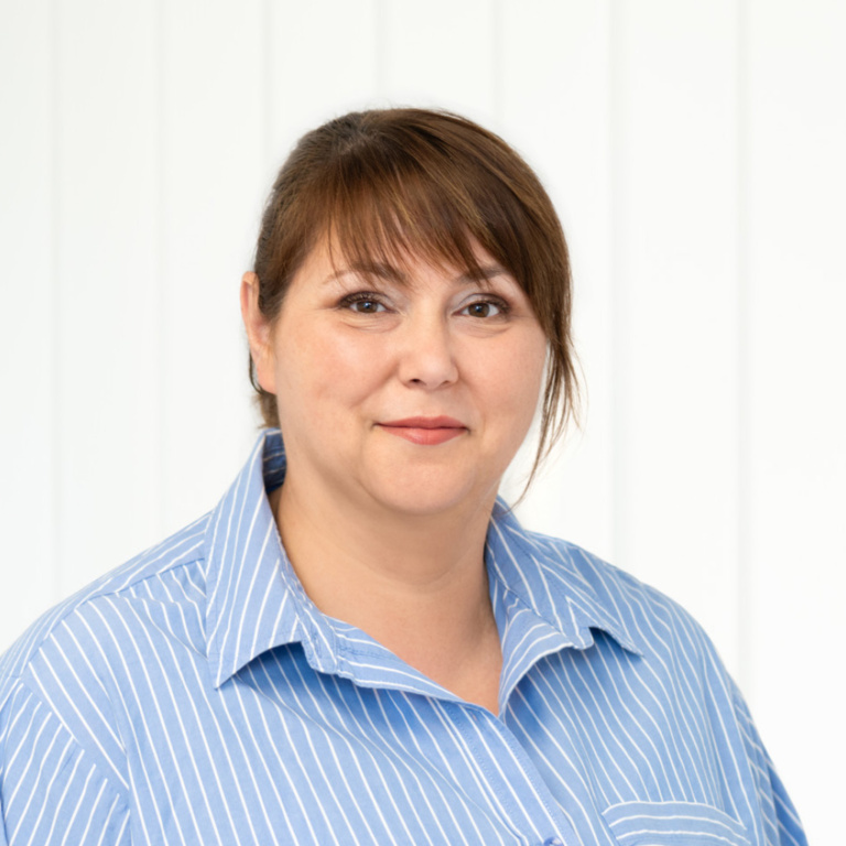 Frau mit braunen Haaren und blauer gestreifter Bluse vor einem hellen Hintergrund, lächelnd und freundlich wirkend.