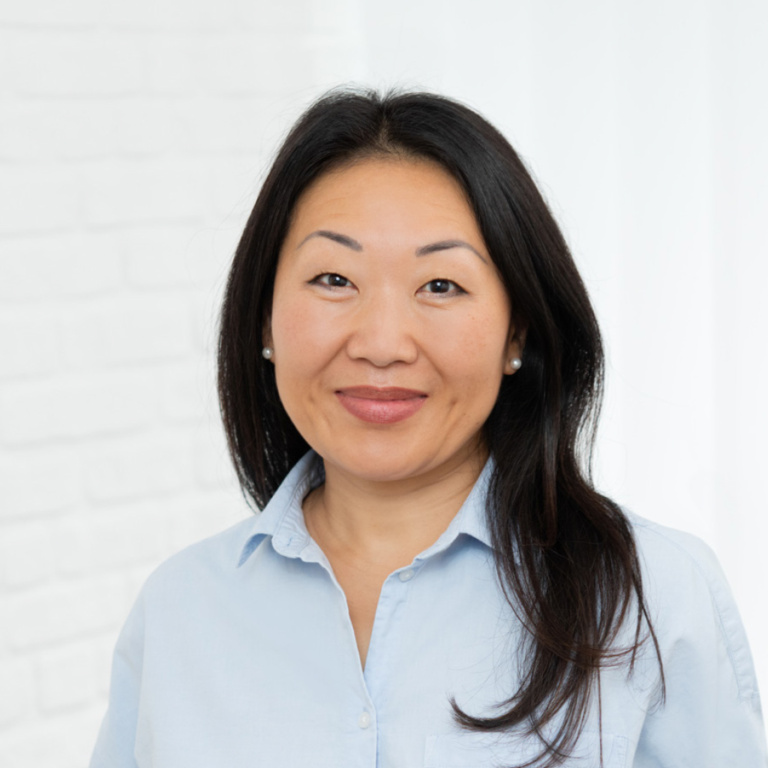 Porträt einer lächelnden Frau mit langen, dunklen Haaren, die ein hellblaues Hemd trägt.