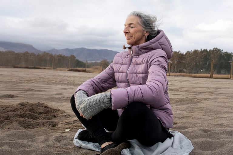 Eine ältere Frau sitzt meditierend auf einem Sandstrand, trägt eine lila Daunenjacke und graue Handschuhe. Im Hintergrund sind sanfte Hügel und bewaldete Flächen zu sehen, während der Himmel bewölkt ist.