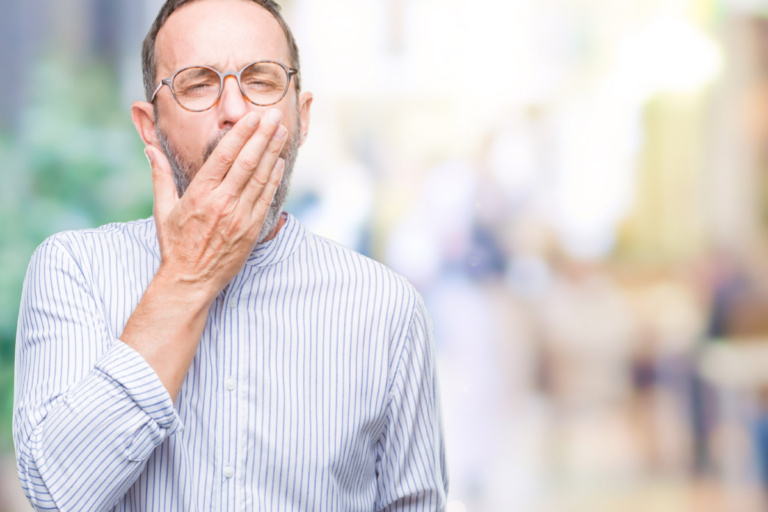Ein Mann mit Brille und Bart trägt ein gestreiftes Hemd und gähnt, während er in einem hellen, unscharfen Hintergrund steht.