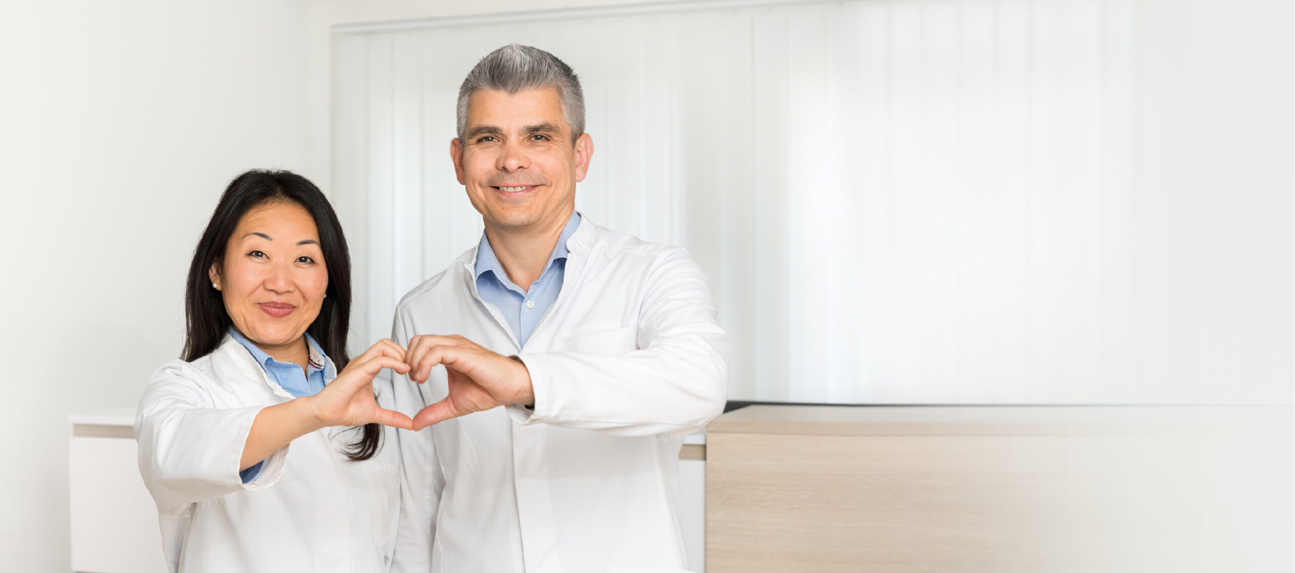 Ärzteteam zeigt Herzsymbol mit Händen in einer modernen Praxis, Ausdruck von Empathie und Patientenorientierung.