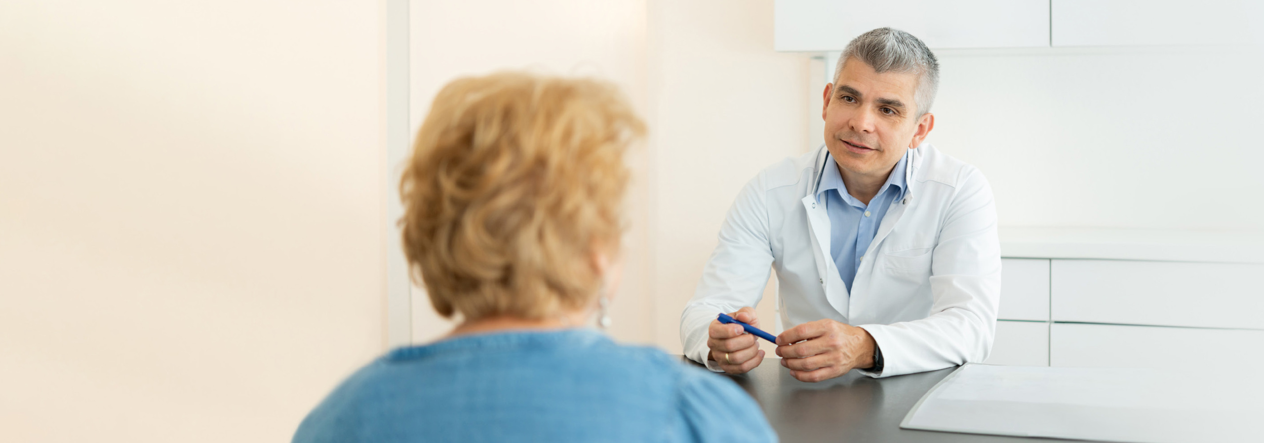 Ein Arzt führt ein Gespräch mit einer Patientin in einer modernen Arztpraxis. Der Arzt trägt einen weißen Kittel und hält einen Stift in der Hand, während die Patientin ihm aufmerksam zuhört.