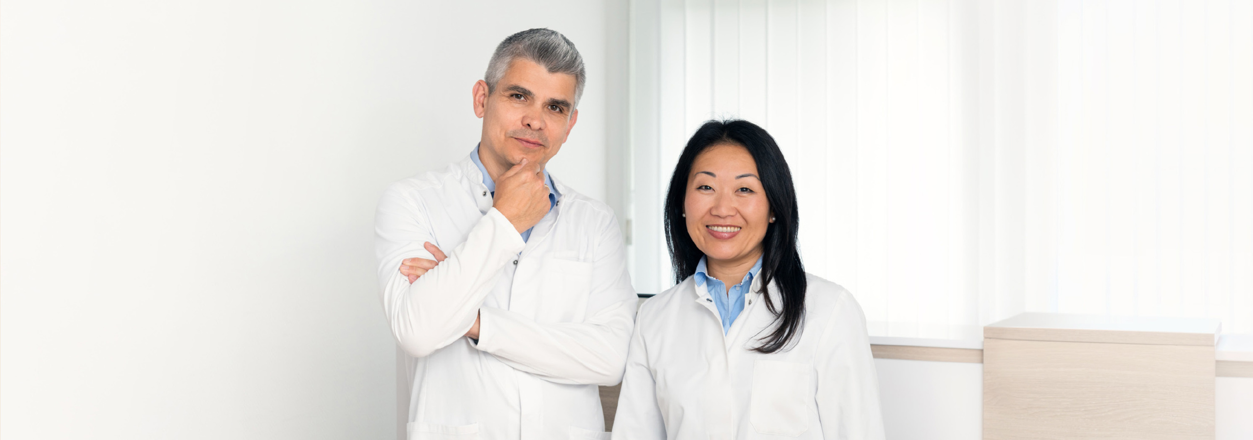 Ein Arzt und eine Ärztin in weißen Kitteln stehen in einem modernen Behandlungsraum und lächeln freundlich in die Kamera.