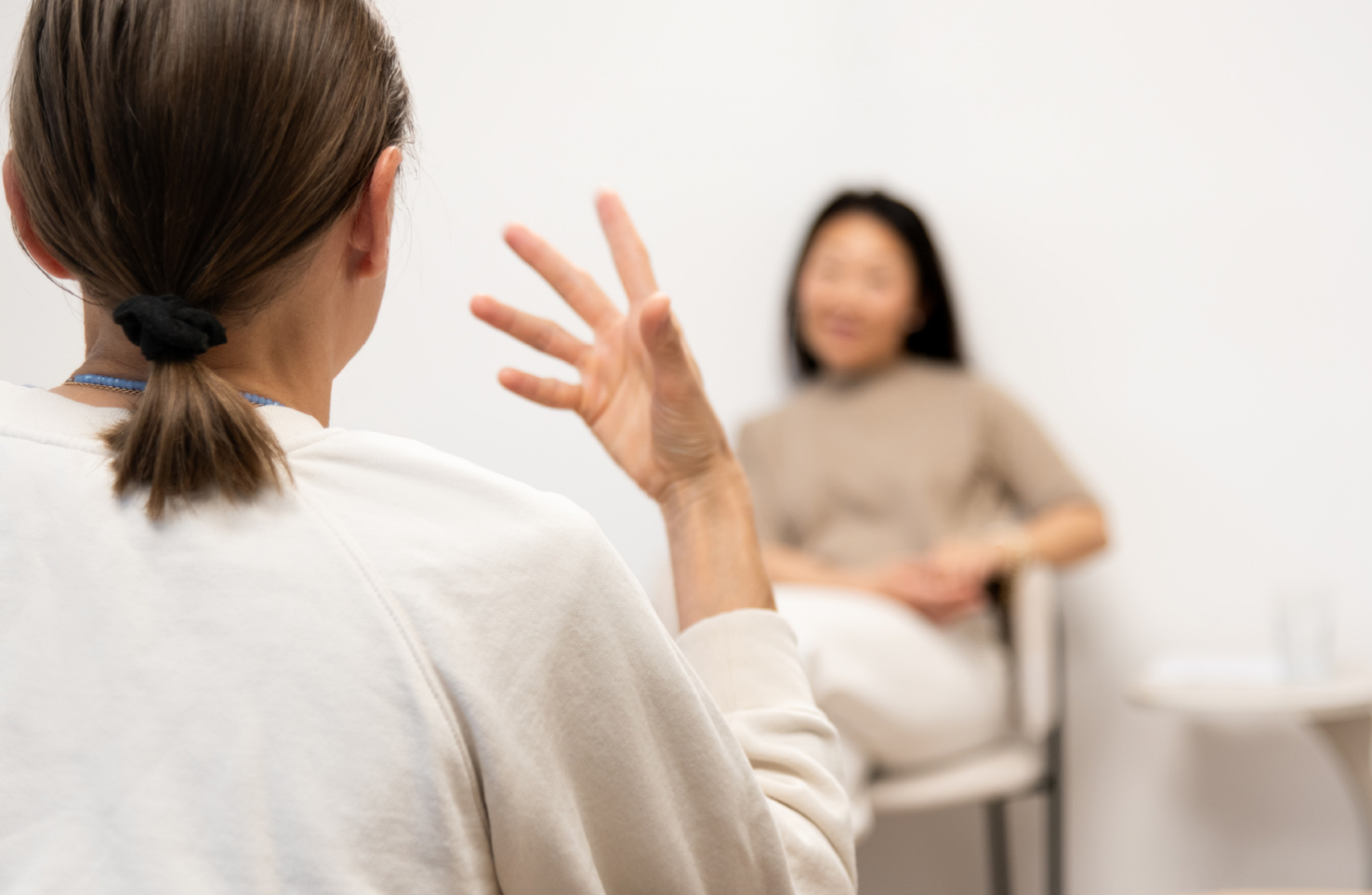 Eine Person mit zurückgebundenem Haar spricht gestikulierend mit einer anderen Person, die entspannt auf einem Stuhl sitzt. Der Hintergrund ist neutral und hell, was die Gesprächsatmosphäre unterstreicht.