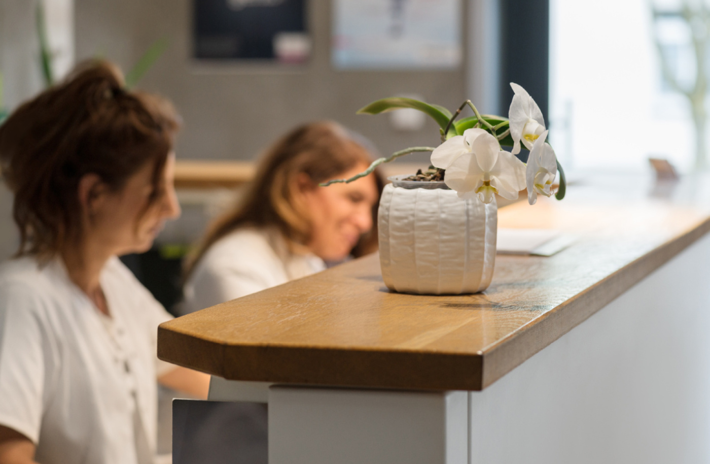 Blütenweiße Orchidee in einer eleganten Vase auf einem Empfangstresen, im Hintergrund zwei Mitarbeiterinnen in weißen Oberteilen, die an der Rezeption arbeiten.