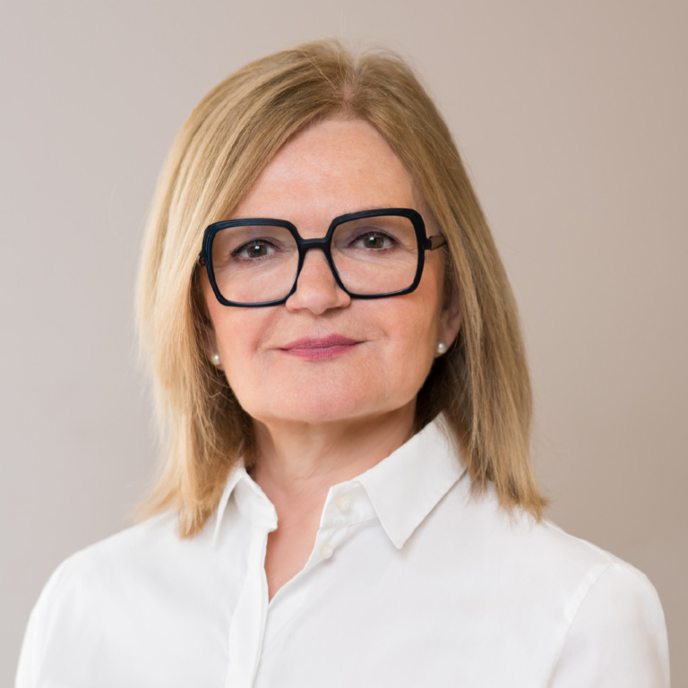 Frau mit blonden Haaren, die eine weiße Bluse trägt und eine Brille mit dicken Rahmen aufhat.