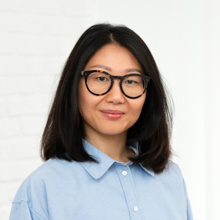 Frau mit schwarzen, runden Brillen und mittellangen, glatten Haaren trägt ein hellblaues Hemd.