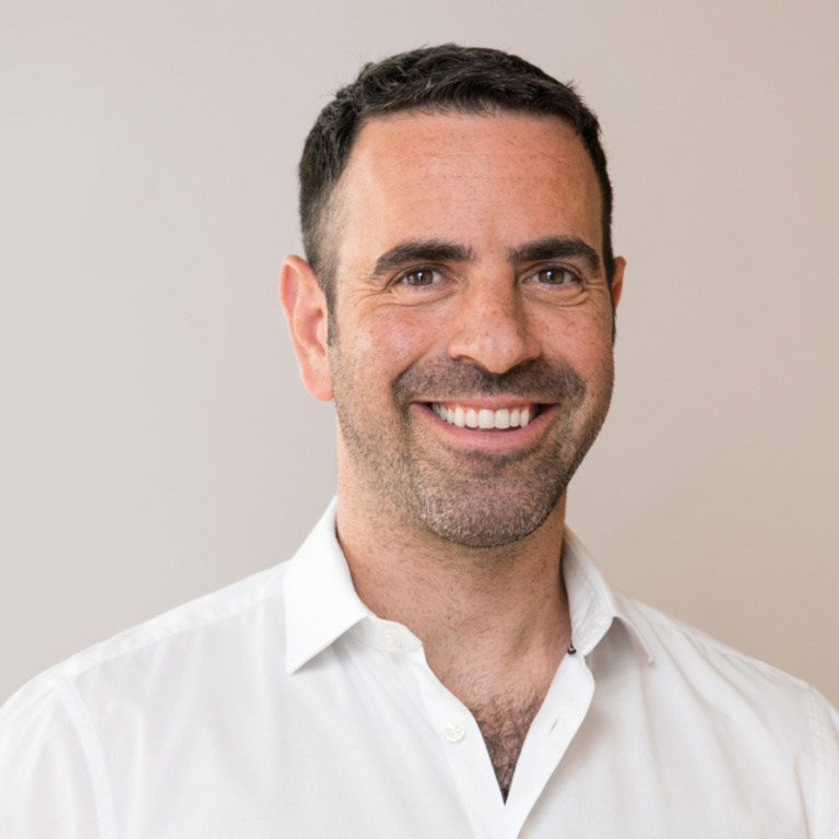 Ein lächelnder Mann mit kurzen, dunklen Haaren trägt ein weißes Hemd.